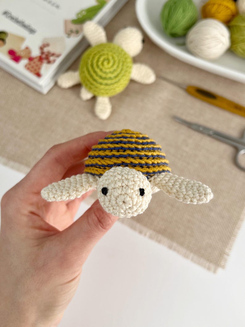 Puede incluir: Una tortuga de ganchillo hecha a mano con un caparaz&oacute;n a rayas amarillas y azul marino, cabeza y extremidades color crema y ojos negros. Otra tortuga est&aacute; en el fondo, junto con hilo y material de manualidades. Un art&iacute;culo artesanal.