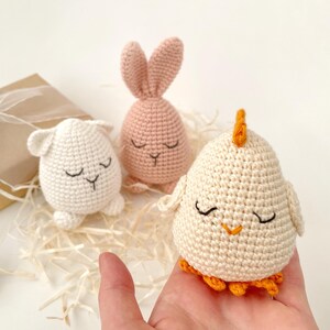 May include: Three crocheted stuffed animals: a pink bunny, a white lamb, and a white chick with an orange beak and feet. The animals are all sitting on a bed of shredded paper.