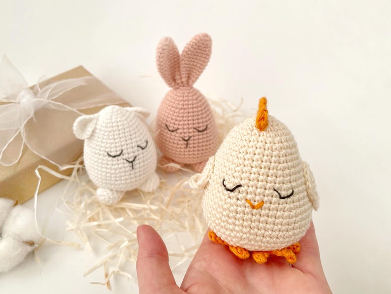 May include: Three crocheted stuffed animals: a pink bunny, a white lamb, and a white chick with an orange beak and feet. The animals are all sitting on a bed of shredded paper.