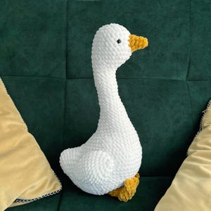 May include: A white, plush goose toy with a long neck and yellow beak and feet. The stuffed animal is made of a soft, textured material and is sitting on a green surface.