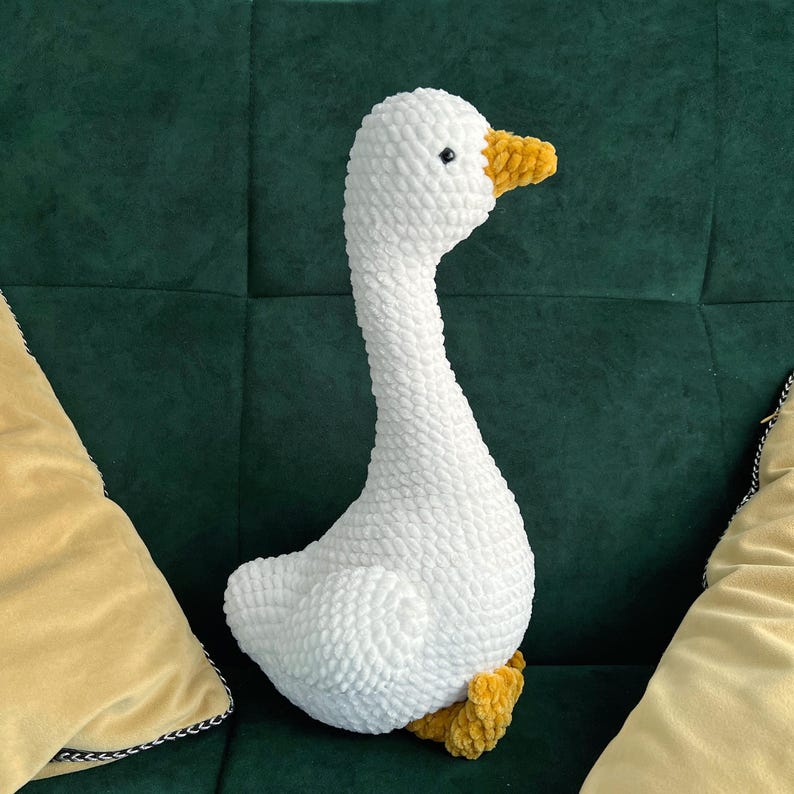 May include: A white, plush goose toy with a long neck and yellow beak and feet. The stuffed animal is made of a soft, textured material and is sitting on a green surface.