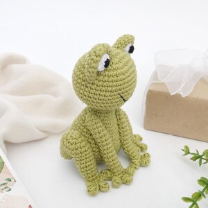 Op de afbeelding: Een handgemaakte, lichtgroene gehaakte kikker met zwarte en witte ogen en een lachende mond. De kikker zit op een wit oppervlak. Een cadeauverpakking verpakt in bruin papier en vastgebonden met een wit lint staat op de achtergrond.