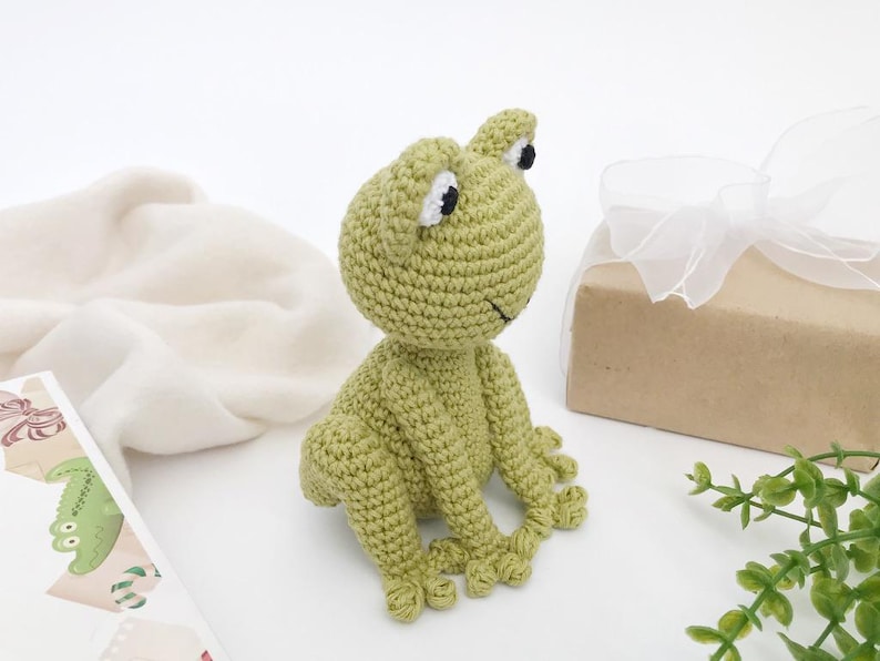 Op de afbeelding: Een handgemaakte, lichtgroene gehaakte kikker met zwarte en witte ogen en een lachende mond. De kikker zit op een wit oppervlak. Een cadeauverpakking verpakt in bruin papier en vastgebonden met een wit lint staat op de achtergrond.