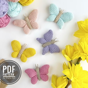 Może przedstawiać: Pięć wzorów na szydełkowe amigurumki w kształcie motyli w różnych kolorach, w tym różowym, żółtym, fioletowym, niebieskim i jasnoniebieskim. Motyle są ułożone na białym tle z żółtymi kwiatami w tle. Tekst "PDF PATTERN IN ENGLISH" jest wyświetlany na brązowym pudełku.