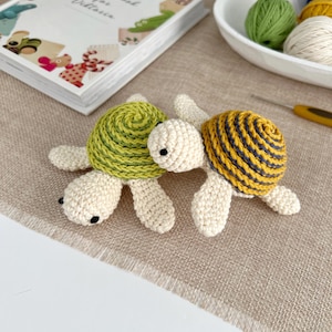 Puede incluir: Dos tortugas de ganchillo. Una tiene un caparaz&oacute;n verde, la otra un caparaz&oacute;n amarillo con rayas azules. Ambas tienen cuerpos blanquecinos. Est&aacute;n sobre una superficie beige. Un libro y lana en el fondo.