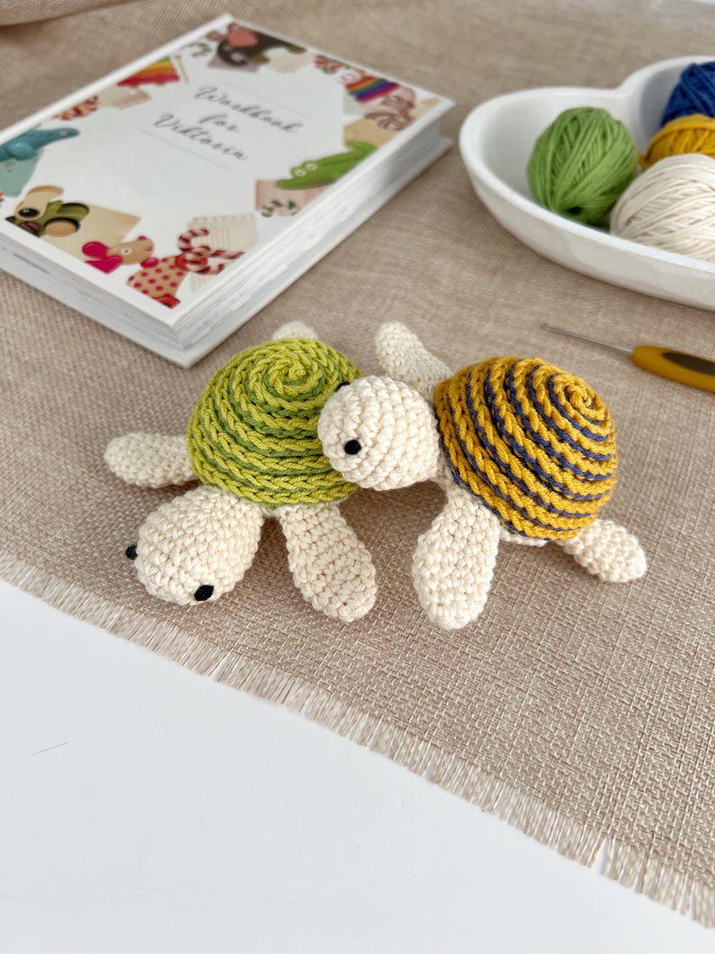 Puede incluir: Dos tortugas de ganchillo. Una tiene un caparaz&oacute;n verde, la otra un caparaz&oacute;n amarillo con rayas azules. Ambas tienen cuerpos blanquecinos. Est&aacute;n sobre una superficie beige. Un libro y lana en el fondo.