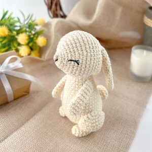 May include: A crocheted white bunny with closed eyes and a pink nose. The bunny is standing on a light brown surface.