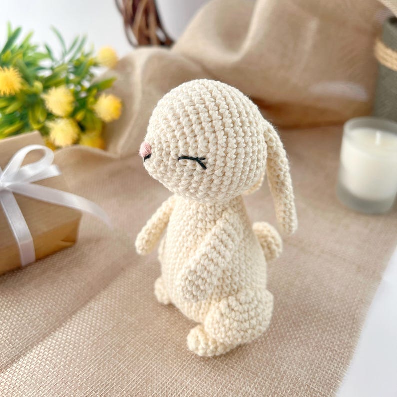 May include: A crocheted white bunny with closed eyes and a pink nose. The bunny is standing on a light brown surface.
