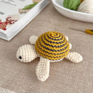 Puede incluir: Una tortuga de ganchillo hecha a mano con un caparaz&oacute;n amarillo y azul marino y un cuerpo color crema. La tortuga est&aacute; sobre una superficie beige. Un libro y lana est&aacute;n en el fondo.