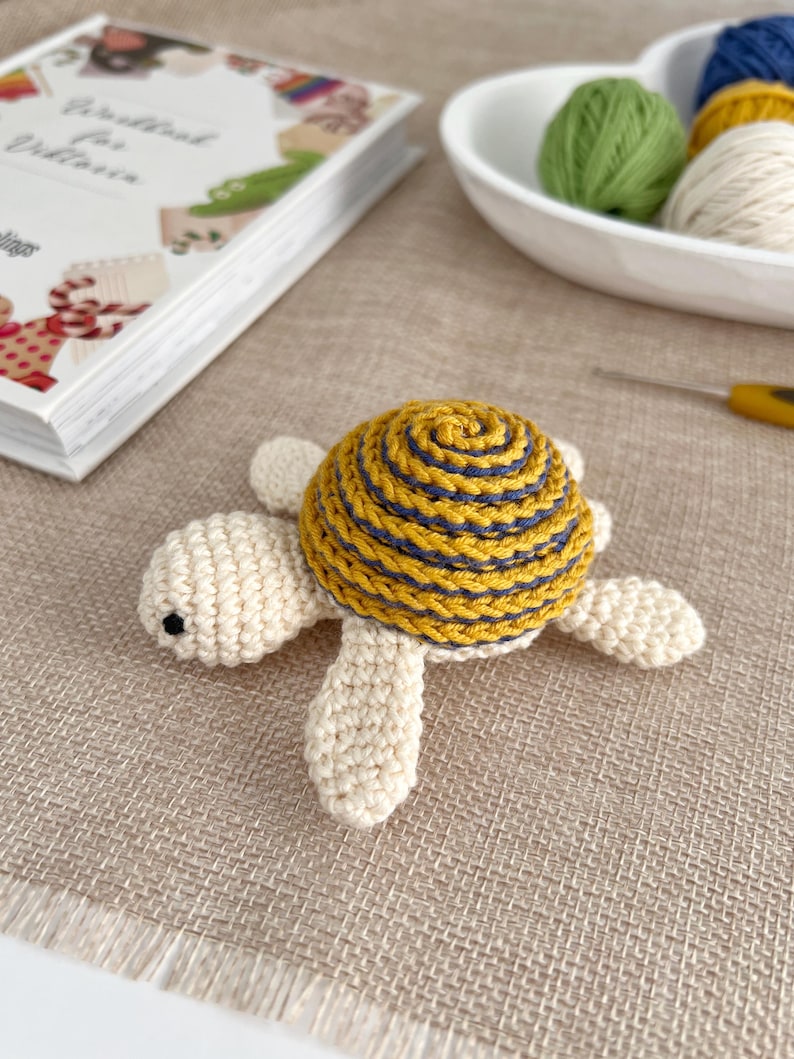 Puede incluir: Una tortuga de ganchillo hecha a mano con un caparaz&oacute;n amarillo y azul marino y un cuerpo color crema. La tortuga est&aacute; sobre una superficie beige. Un libro y lana est&aacute;n en el fondo.