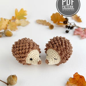Peut inclure: Deux petits hérissons au crochet avec un dos texturé marron et un visage crème. Un hérisson a des yeux noirs ouverts, tandis que l'autre a les yeux fermés. Un badge circulaire dans le coin supérieur droit contient le texte PDF PATTERN IN ENGLISH.