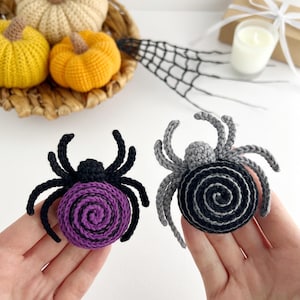 May include: Two crocheted spider ornaments. One spider has a purple spiral body with black legs and head. The other spider has a black and gray spiral body with gray legs and head. Both spiders feature eight legs and a round, textured body.