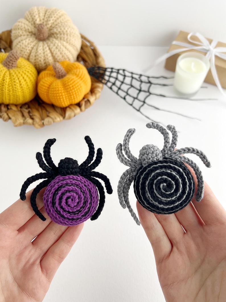 May include: Two crocheted spider ornaments. One spider has a purple spiral body with black legs and head. The other spider has a black and gray spiral body with gray legs and head. Both spiders feature eight legs and a round, textured body.