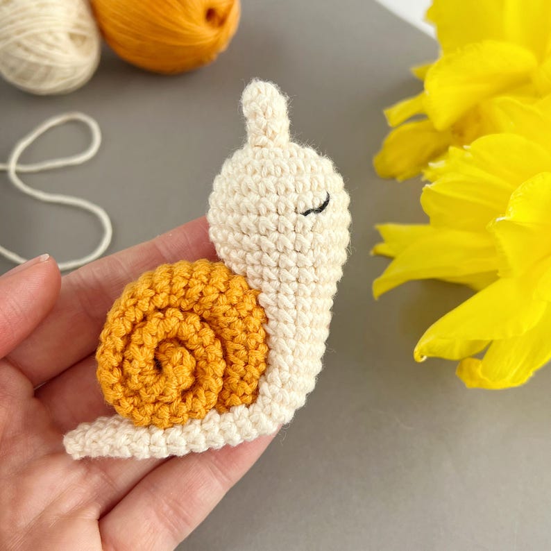 Pode incluir: Um brinquedo de amigurumi de caracol de croch&ecirc; com um corpo branco e uma concha amarela. O caracol tem olhos pretos e uma pequena boca.