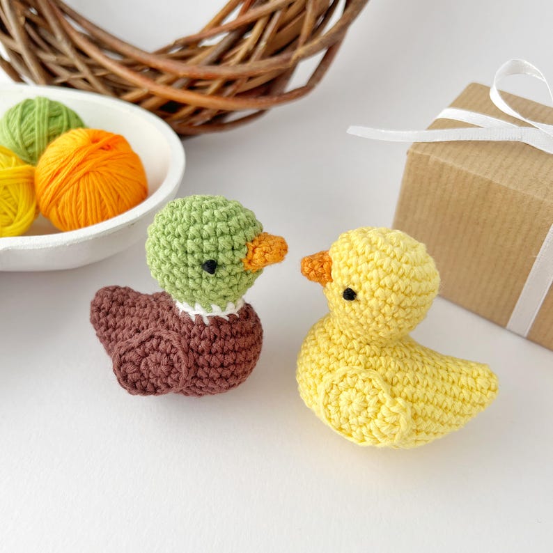 May include: Two handmade crochet ducklings. One duckling is yellow with an orange beak, and the other is brown with a green head and orange beak. A white bowl with yellow, green, and orange yarn balls is in the background.