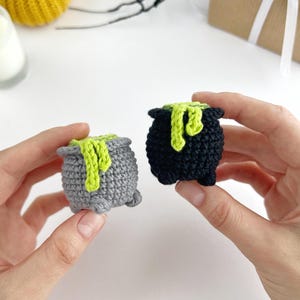 May include: Two small crocheted cauldrons, one in grey and one in black, each featuring a textured surface and a bright lime green yarn accent draped over the rim to resemble bubbling liquid, with small feet at the base of each vessel.