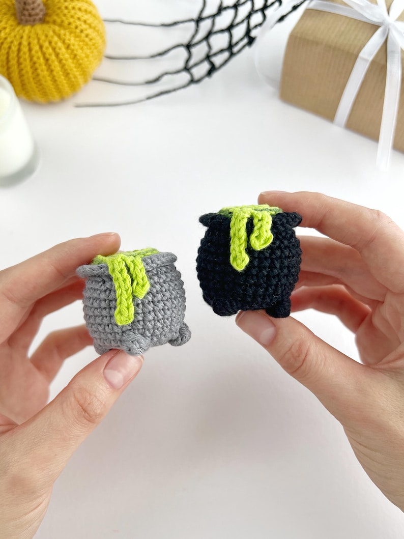 May include: Two small crocheted cauldrons, one in grey and one in black, each featuring a textured surface and a bright lime green yarn accent draped over the rim to resemble bubbling liquid, with small feet at the base of each vessel.