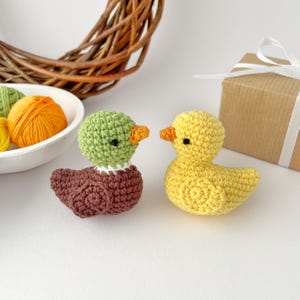 May include: Two crocheted ducklings. One duckling is yellow with an orange beak and black eyes. The other duckling has a green head, brown body, and orange beak. A white bowl with orange and green yarn is in the background.