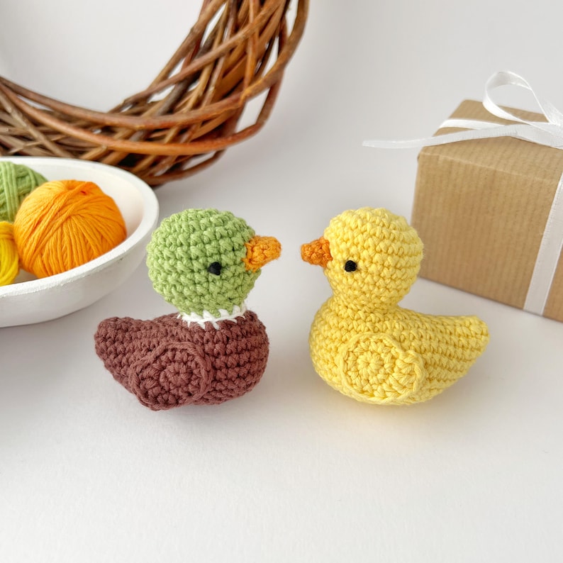 May include: Two crocheted ducklings. One duckling is yellow with an orange beak and black eyes. The other duckling has a green head, brown body, and orange beak. A white bowl with orange and green yarn is in the background.