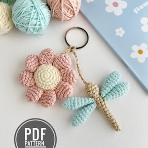 May include: A handmade crochet keychain featuring a pink flower and a dragonfly with blue wings. The keychain is attached to a metal ring. The image also shows balls of yarn in various pastel colors and a blue notebook.