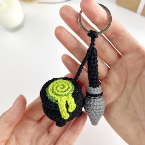 May include: A crochet keychain featuring a black cauldron with a swirling lime green potion spilling over the rim, attached to a silver metal ring. Next to the cauldron hangs a small, grey and black crochet wand with a pointed tip.