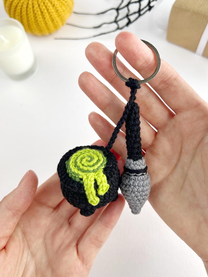 May include: A crochet keychain featuring a black cauldron with a swirling lime green potion spilling over the rim, attached to a silver metal ring. Next to the cauldron hangs a small, grey and black crochet wand with a pointed tip.