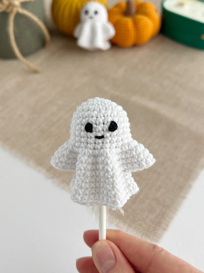 May include: A small white crocheted ghost figure with black embroidered eyes and a simple mouth, attached to a thin white stick. The ghost has a rounded head and draped arms, creating a soft, textured appearance for this handmade decorative item.