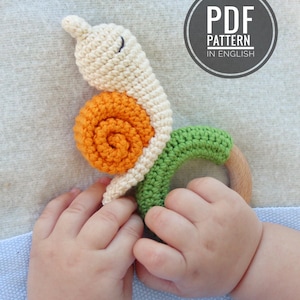 May include: A handmade crochet snail baby toy in cream, orange, and green with a wooden ring. The toy is held by a baby's hands. The image includes a circular graphic that says "PDF PATTERN IN ENGLISH".
