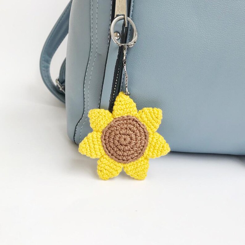 May include: A yellow and brown crochet sunflower charm hanging from a silver-colored keychain. The sunflower has a brown center and yellow petals. The charm is attached to a light blue purse.