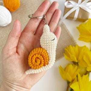 Pode incluir: Um chaveiro de caracol de croch&ecirc; com um corpo branco e uma concha amarela. O chaveiro est&aacute; preso a um chaveiro de prata.