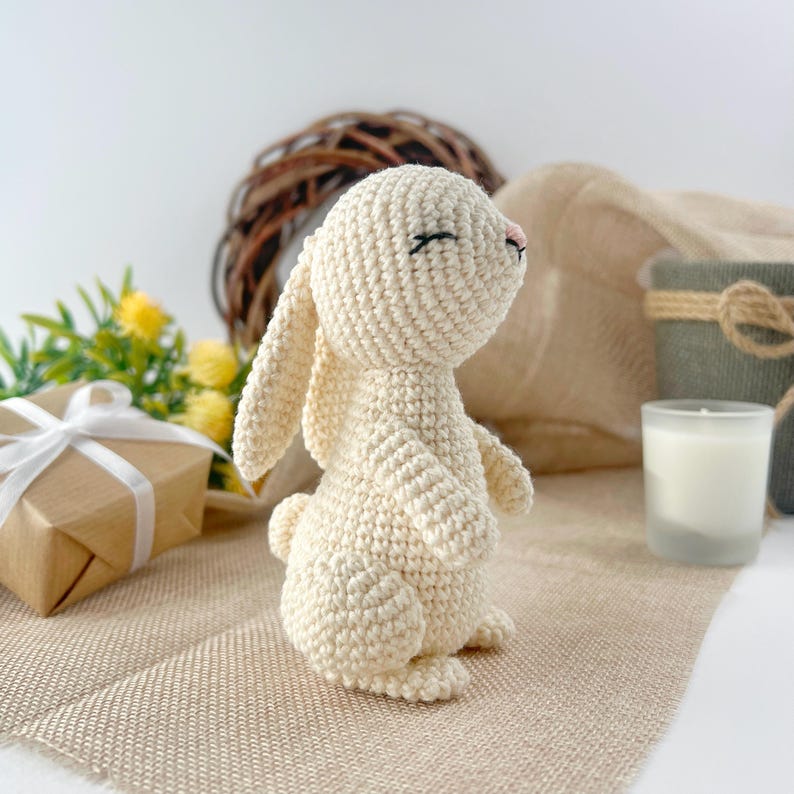 May include: A crocheted white bunny rabbit with closed eyes and a pink nose. The bunny is sitting on a beige burlap surface.