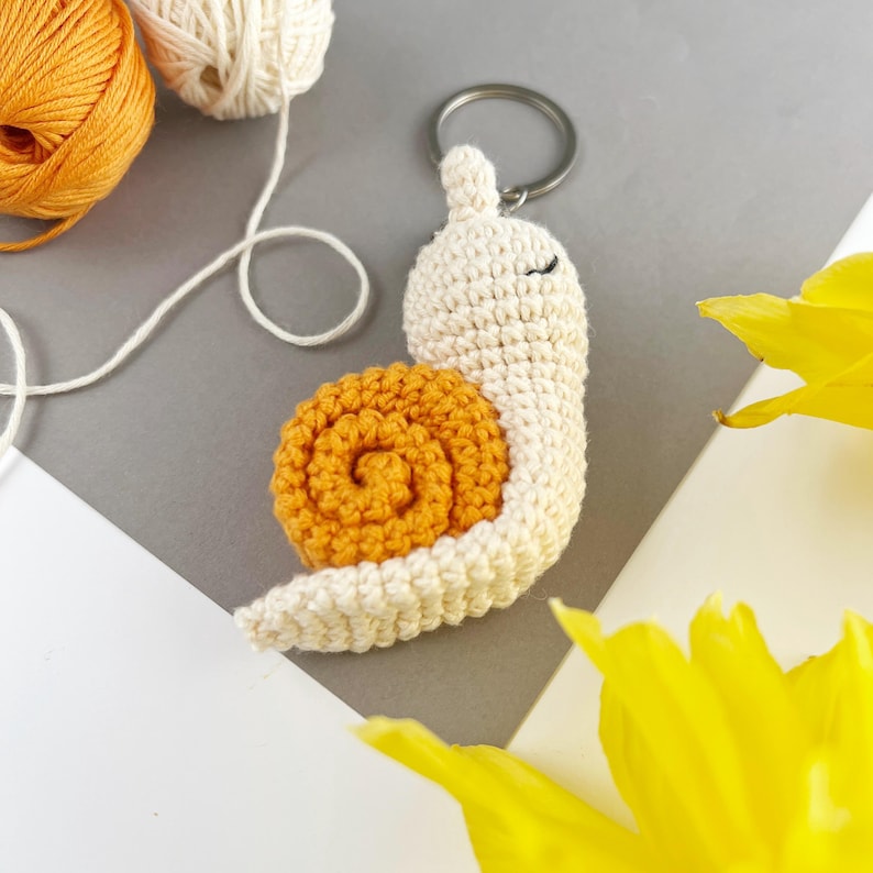 Pode incluir: Um chaveiro de caracol de croch&ecirc; com uma concha branca e uma espiral laranja. O caracol tem olhos pretos e um anel de chave preso.