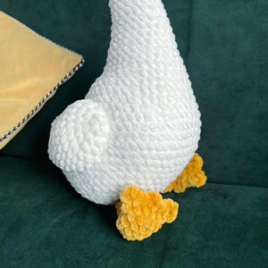 May include: A plush white goose with yellow feet. The goose is made of a soft, textured material. The goose is sitting on a dark green surface. A yellow pillow with a black and white trim is in the background.