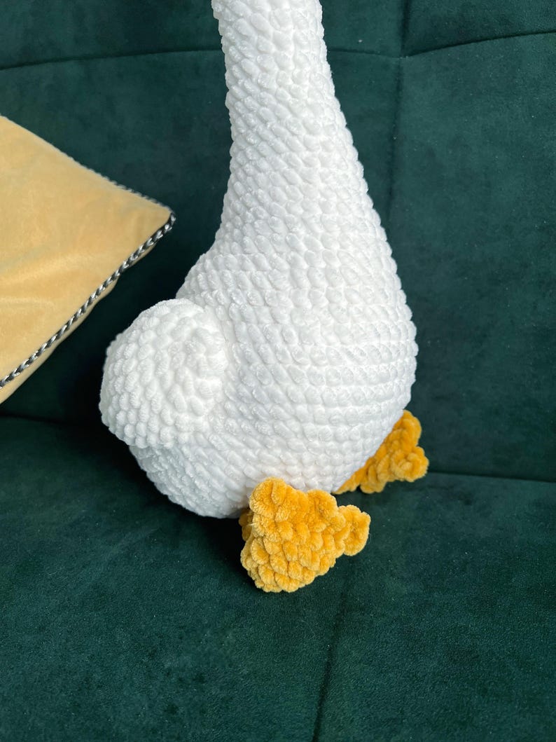 May include: A plush white goose with yellow feet. The goose is made of a soft, textured material. The goose is sitting on a dark green surface. A yellow pillow with a black and white trim is in the background.