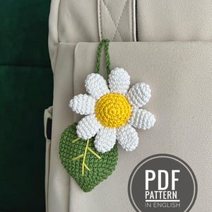 Può includere: Un fiore di margherita all'uncinetto con petali bianchi e un centro giallo, attaccato a una foglia verde. La margherita è appesa a uno zaino beige. L'immagine include una grafica circolare che dice "PDF PATTERN IN ENGLISH".