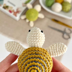 Puede incluir: Tortuga de ganchillo hecha a mano con cabeza y extremidades de color crema, y caparaz&oacute;n amarillo con detalles en espiral azul. La tortuga se sostiene frente a un fondo borroso con otros materiales de artesan&iacute;a y un peque&ntilde;o libro.