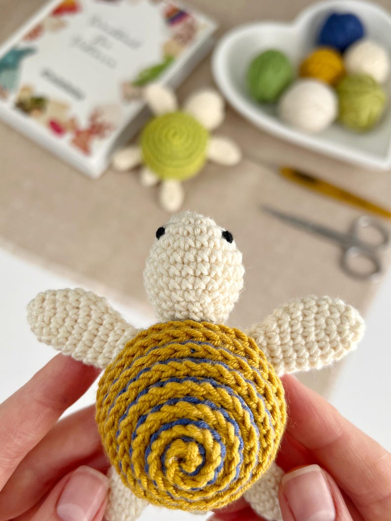 Puede incluir: Tortuga de ganchillo hecha a mano con cabeza y extremidades de color crema, y caparaz&oacute;n amarillo con detalles en espiral azul. La tortuga se sostiene frente a un fondo borroso con otros materiales de artesan&iacute;a y un peque&ntilde;o libro.