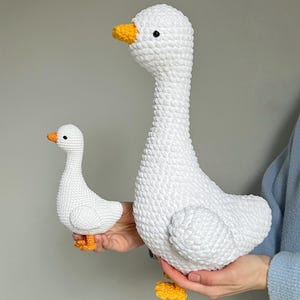 May include: Two white crochet geese, one large and one small, are held in a person's hands. The geese have yellow beaks and feet, and black eyes. The larger goose is approximately 12 inches tall.