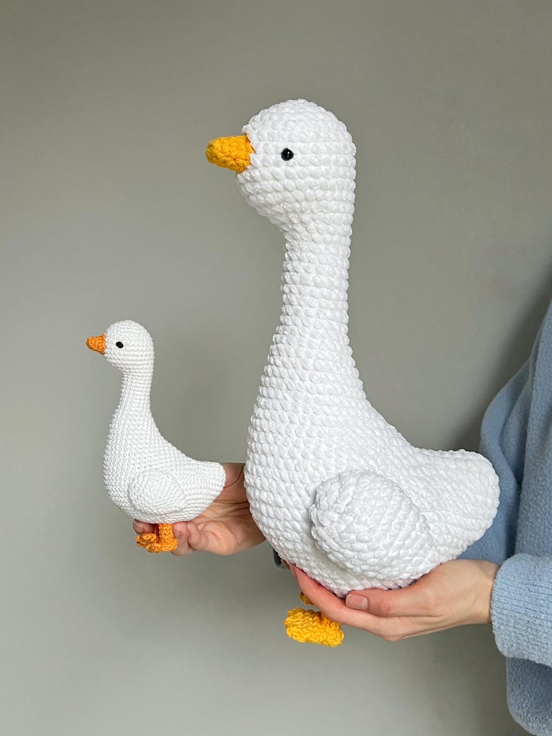 May include: Two white crochet geese, one large and one small, are held in a person's hands. The geese have yellow beaks and feet, and black eyes. The larger goose is approximately 12 inches tall.