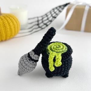 May include: A black crocheted cauldron with a lime green swirled liquid overflowing from the top, accompanied by a small crocheted broomstick featuring a grey bristle end and a black handle, both crafted with textured yarn stitches.