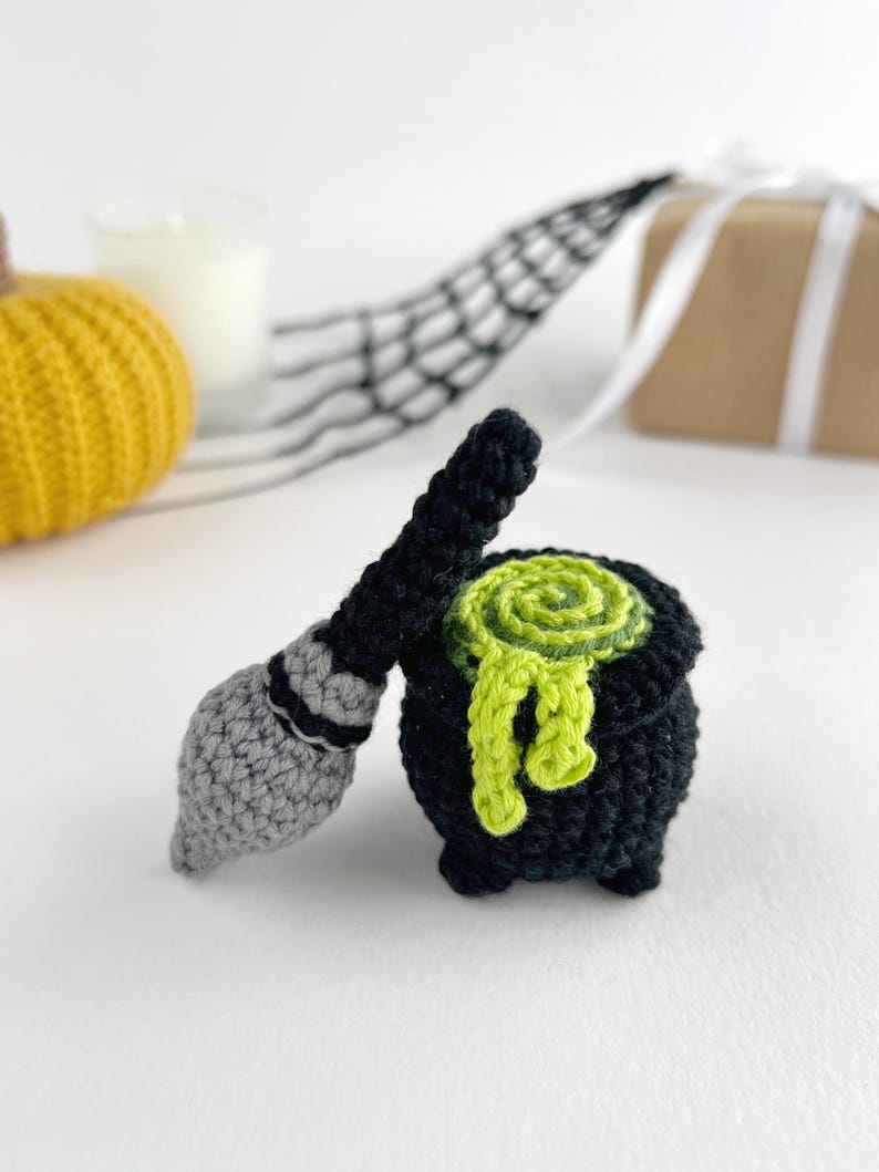 May include: A black crocheted cauldron with a lime green swirled liquid overflowing from the top, accompanied by a small crocheted broomstick featuring a grey bristle end and a black handle, both crafted with textured yarn stitches.