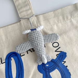 May include: A crocheted gray airplane charm with white propeller and tail accents is attached to a canvas tote bag. The bag has blue abstract lettering. The airplane is suspended by a gray string.