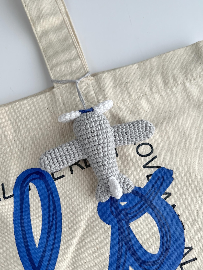 May include: A crocheted gray airplane charm with white propeller and tail accents is attached to a canvas tote bag. The bag has blue abstract lettering. The airplane is suspended by a gray string.