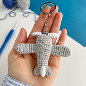 May include: A crocheted airplane keychain in gray and white, with a silver keyring. The wings and tail are detailed, and the keychain is held in a hand. The background includes blue and white yarn and a crochet hook.