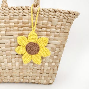 May include: A woven straw tote bag with a yellow crochet sunflower charm. The sunflower has a brown center and yellow petals. The bag is made of natural-colored straw and has a woven design.