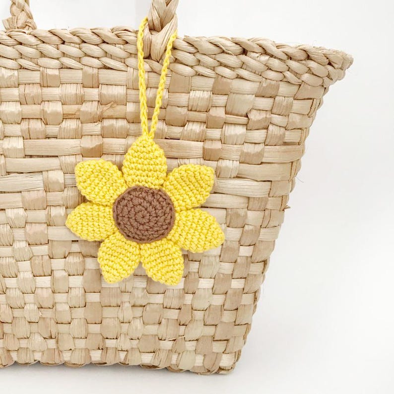 May include: A woven straw tote bag with a yellow crochet sunflower charm. The sunflower has a brown center and yellow petals. The bag is made of natural-colored straw and has a woven design.