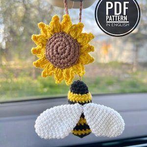 May include: Crocheted sunflower and bee car charm. The sunflower is yellow with a brown center, and the bee has a black and yellow body with white wings. The charm hangs from a brown string. The image includes a PDF pattern in English.