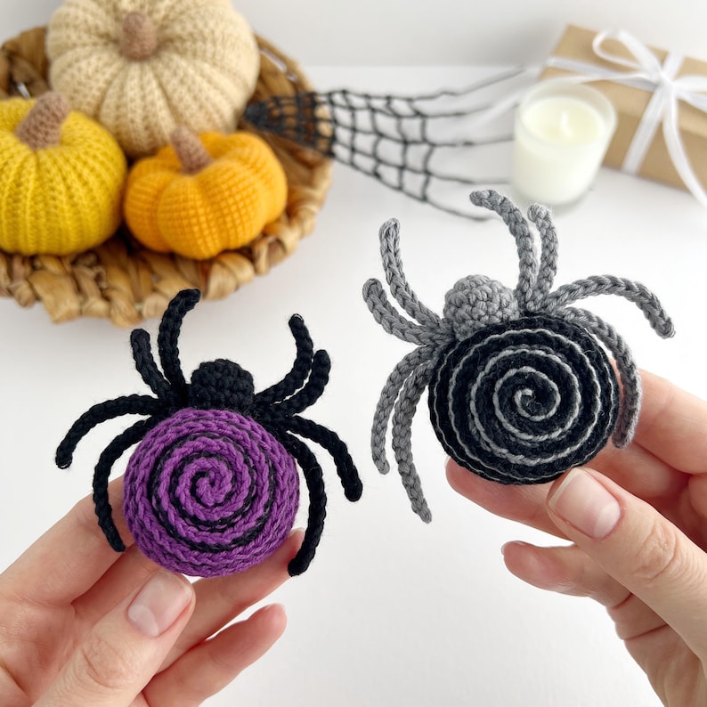 May include: Two handmade crochet spiders. One spider features a purple and black spiral body with black legs. The second spider has a grey and black spiral body with grey legs. Both spiders have eight legs and a small round head attached to the body.