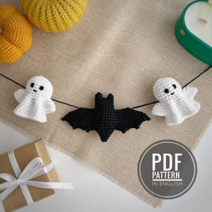 Könnte beinhalten: Eine Halloween-Girlande aus Häkelarbeit mit einer schwarzen Fledermaus und zwei weißen Geistern. Die Girlande ist an einem schwarzen Faden aufgereiht und auf einer beigen Oberfläche präsentiert. Das Bild enthält auch zwei gelbe gehäkelte Kürbisse und eine Geschenkbox.