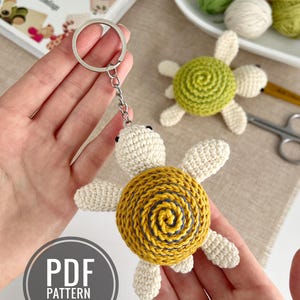 Puede incluir: Llavero de tortuga de ganchillo con caparaz&oacute;n amarillo y azul, cabeza y extremidades color crema y un llavero plateado. La imagen tambi&eacute;n muestra un patr&oacute;n PDF en ingl&eacute;s y otras tortugas de ganchillo.